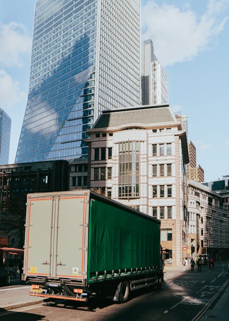 A large green commercial moving truck with closed rear doors is parked on a city street next to modern glass skyscrapers and older stone buildings. The truck is positioned adjacent to the curb, ready