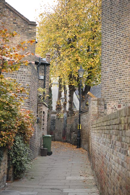 The image depicts a narrow residential street in Clapham with a wet asphalt road flanked by colorful houses on the right and a brick wall with small garages on the left. The houses are painted in past